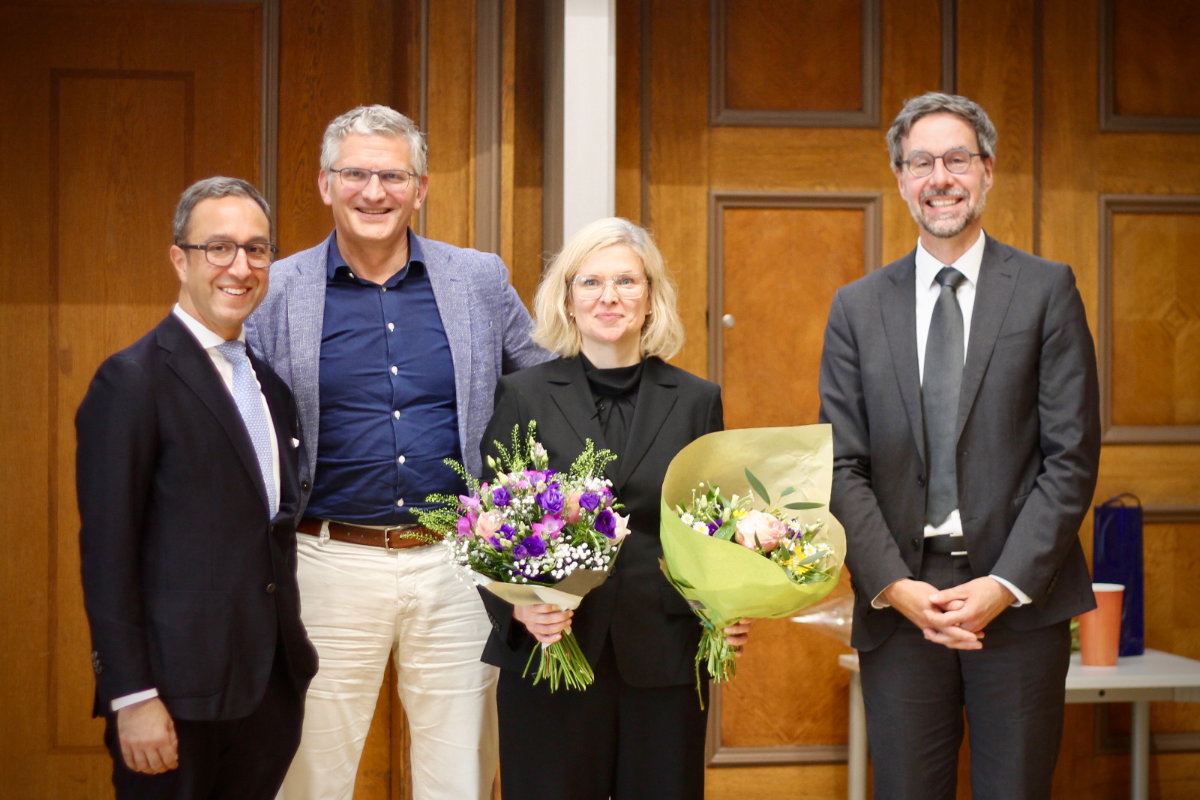 Vier Personen in formeller Kleidung stehen vor einer Holzwand, zwei halten Blumensträuße
