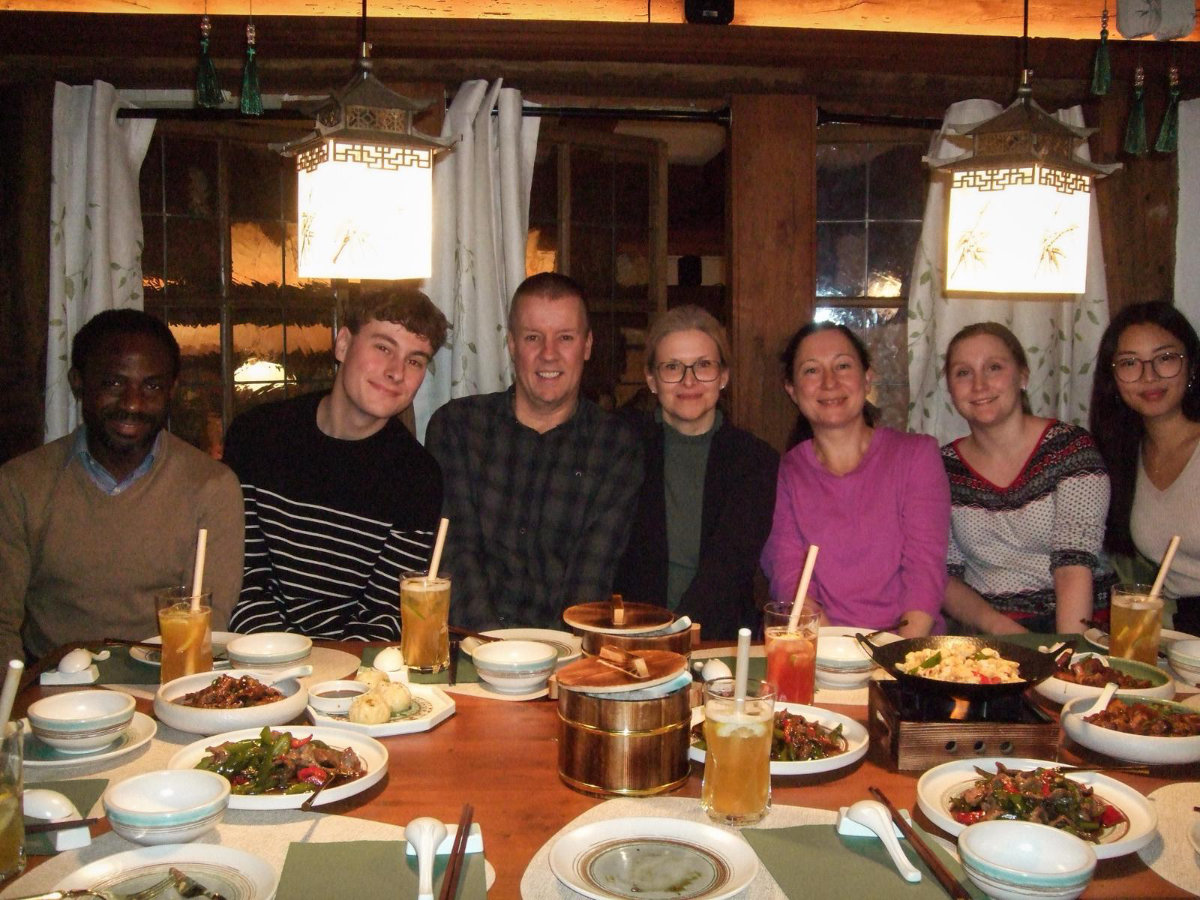 Gruppe von sechs Personen sitzt an einem Tisch mit asiatisch anmutenden Speisen und Getränken in einem Restaurant mit hölzernen Wänden und dekorativen Lampen
