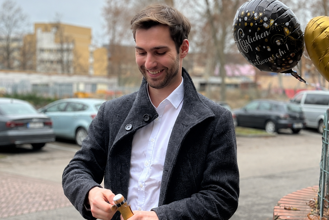 Lächelnder Mann in dunklem Mantel öffnet eine Sektflasche im Freien neben einem Korb mit Blumen und zwei Luftballons