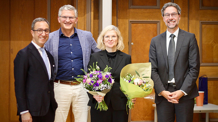 Vier Personen in formeller Kleidung stehen vor einer Holzwand, zwei halten Blumensträuße