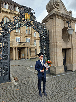Person in Anzug mit Doktorhut hält Blumenstrauß und Glas vor einem historischen Torbogen aus Stein und Schmiedeeisen