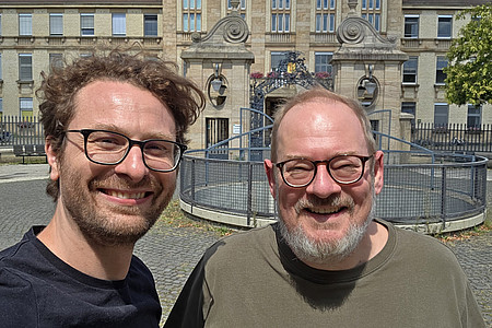 Zwei Männer mit Brille lächeln in die Kamera vor einem historischen Gebäude mit Rundbrunnen.
