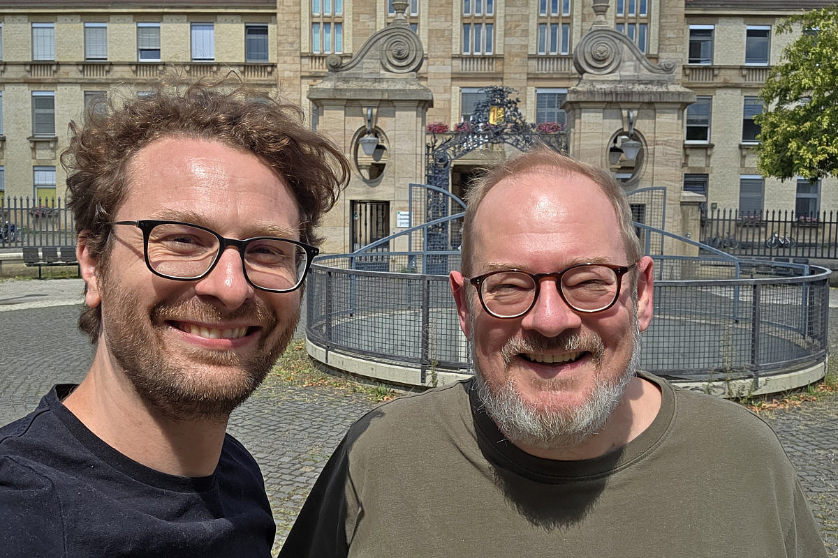 Zwei Männer mit Brille lächeln in die Kamera vor einem historischen Gebäude mit Rundbrunnen.